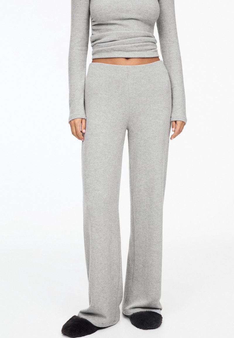 Femme portant un pantalon large en maille gris clair taille haute et un top court à manches longues assorti, associé à des chaussons noirs duveteux.