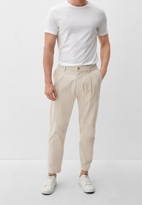Beige plissébroek van lichtgewicht stof, voorzien van een knoopsluiting, taps toelopende pijpen en een casual pasvorm, gecombineerd met witte sneakers.