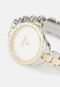Reloj redondo con un dial blanco, bisel estriado en tono dorado, correa de acero inoxidable plateado, acentos dorados y marcadores de hora sencillos.