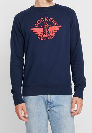 Mężczyzna noszący granatową bluzę z czerwonym logo "Dockers" przedstawiającym kotwicę i skrzydła, w połączeniu z jasnoniebieskimi jeansami, na jednolitym tle.
