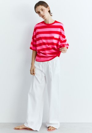 Junge Person trägt ein lockeres rosa-rot gestreiftes Hemd mit weißen, weit geschnittenen Hosen und rosa Sandalen, steht an einer schlichten weißen Wand.