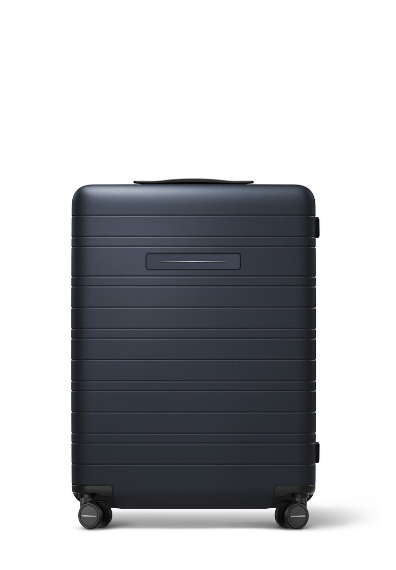 Valise rigide de couleur bleu marine mat, avec des rainures horizontales, une poignée supérieure et quatre roues noires pour plus de mobilité.