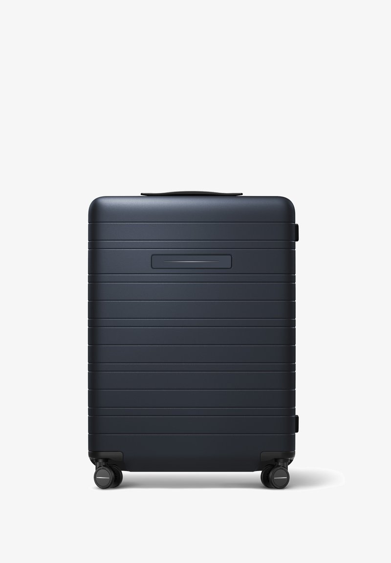 Valise rigide de couleur bleu marine mat, avec des rainures horizontales, une poignée supérieure et quatre roues noires pour plus de mobilité.