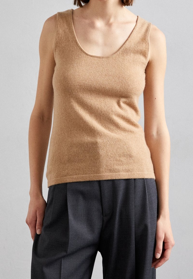 Femme portant un haut en maille sans manches de couleur camel et un pantalon plissé gris foncé, debout devant un fond gris clair.