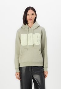 Gaiši zaļš kapucis ar pūkainu balto "UGG" logo, ar regulējamu kapuci un kangurū jaku, izgatavots no mīksta auduma ar ribotām malām un piedurknēm.