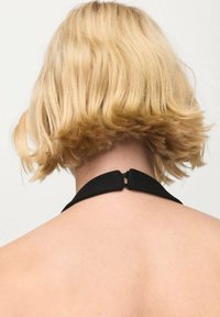 Black halter strap with a metal clasp connects at the nape of a neck featuring blonde, chin-length, wavy hair. Background is neutral.