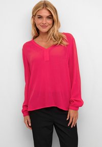 Blusa fucsia de un tejido ligero y transparente con escote en V y mangas largas, que presenta puños fruncidos y un corte relajado. Combinada con pantalones negros.