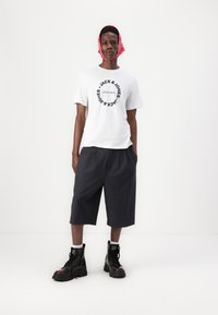 White t-shirt with black circular logo text, dark gray pleated shorts, and black lace-up combat boots with chunky soles. Red bandana around head.