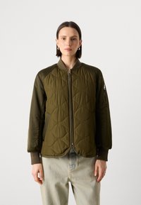 Olivenfarget, quiltet bomberjakke med glidelåsfront, ribbet krage, glidelåslomme på armen og strukturert stoff. Kombinert med lyse denimbukser.