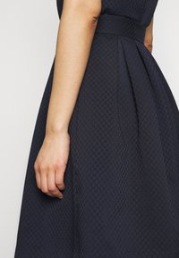 Námořnické modré šaty s texturovaným hexagonálním vzorem, s vypasovaným pasem a plisovanou sukní. Viditelná ruka drží stranu šatů.