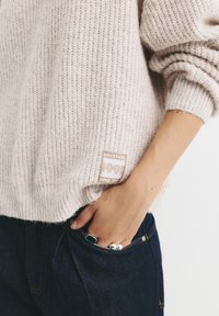 Helle beige Strickpullover mit strukturierter Oberfläche, ausgestattet mit einem seitlich angenähten Etikett. Das Model trägt dunkelblaue Jeans und Ringe.