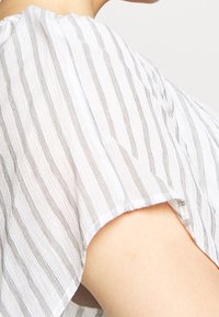 Top blanco con sutiles rayas verticales en gris claro. Textura suave y ligera, ajuste claramente holgado y mangas anchas.