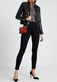 Giacca di pelle nera con zip dorate, top con stampa leopardo, jeans skinny neri, borsa a tracolla rossa e tacchi alti neri.