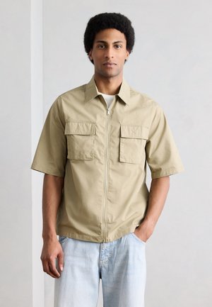 Homme aux cheveux courts et bouclés portant une chemise beige à manches courtes avec fermeture éclair et grandes poches avant, ainsi qu'un jean bleu clair.