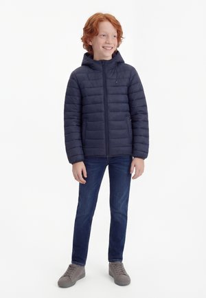 Uśmiechnięty chłopiec z rudymi włosami, ubrany w granatową pikowaną kurtkę z kapturem, niebieskie dżinsy i szare trampki, stojący na białym tle.