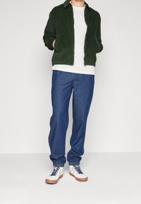 Groene corduroy zip-up jas over een wit t-shirt, gecombineerd met losse blauwe denim jeans en witte sneakers met marineblauwe accenten en gumzolen.
