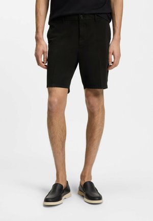 Hombre con pantalones cortos negros a medida y zapatos negros sin cordones con suelas blancas, de pie contra un fondo blanco.