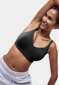 Frau trägt einen schwarzen Sport-BH und graue Jogginghosen, dehnt sich mit einem erhobenen Arm, lächelt, vor einfachem hellem Hintergrund.