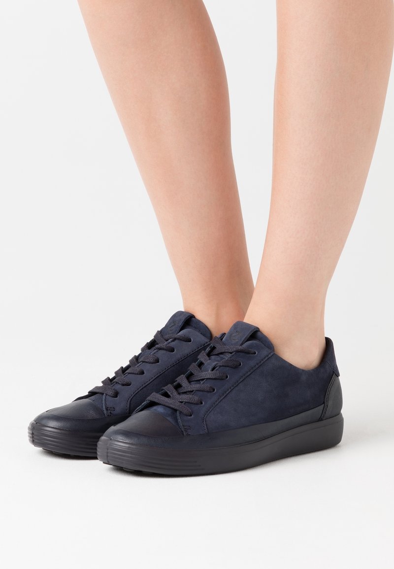 Pieds portant des baskets à lacets bleu marine à coupe basse avec semelles noires sur fond blanc.