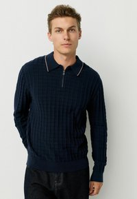 Pulover tricotat bleumarin cu un model texturat în grilaj, guler cu fermoar și garnitură de culoare deschisă contrastantă pe guler, purtat cu blugi întunecați.