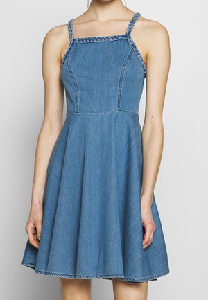 Vrouw met een mouwloze blauwe spijkerjurk met gevlochten bandjes en een uitlopende rok, staand tegen een eenvoudige lichte achtergrond.