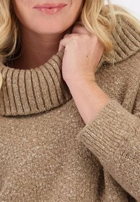 Beige gebreide sweater met een brede, geribbelde col en een textuurstof. Close-up weergave die de zachtheid en handdetails benadrukt.