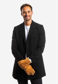 Schwarzer Wollmantel mit Knopfverschluss, getragen über einem weißen Hemd. Braune Lederhandschuhe mit weicher Fütterung, sichtbaren Nähten und Fingeröffnungen.