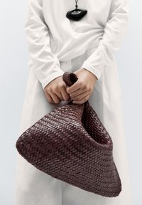 Vävd bordeauxväska med en unik triangulär form, texturerad yta och ett enda, knutet handtag. En enkel ljus klädsel i bakgrunden.