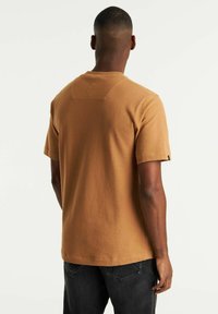 Camicia marrone a maniche corte, con texture, colletto rotondo e dettagli di cucitura discreti sul retro; abbinata a jeans scuri.