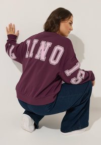 Felpa bordeaux con testo bianco "LINO" e "15" sulle maniche; abbinata a jeans a gamba larga blu scuro e sneakers bianche. Tessuto morbido, vestibilità casual.