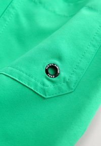 Zelená látka s našitým kapsou a černým kovovým průchodem, na kterém je vyrytý text "NEXT.SWIM 1982". Hladká textura a výrazná barva.