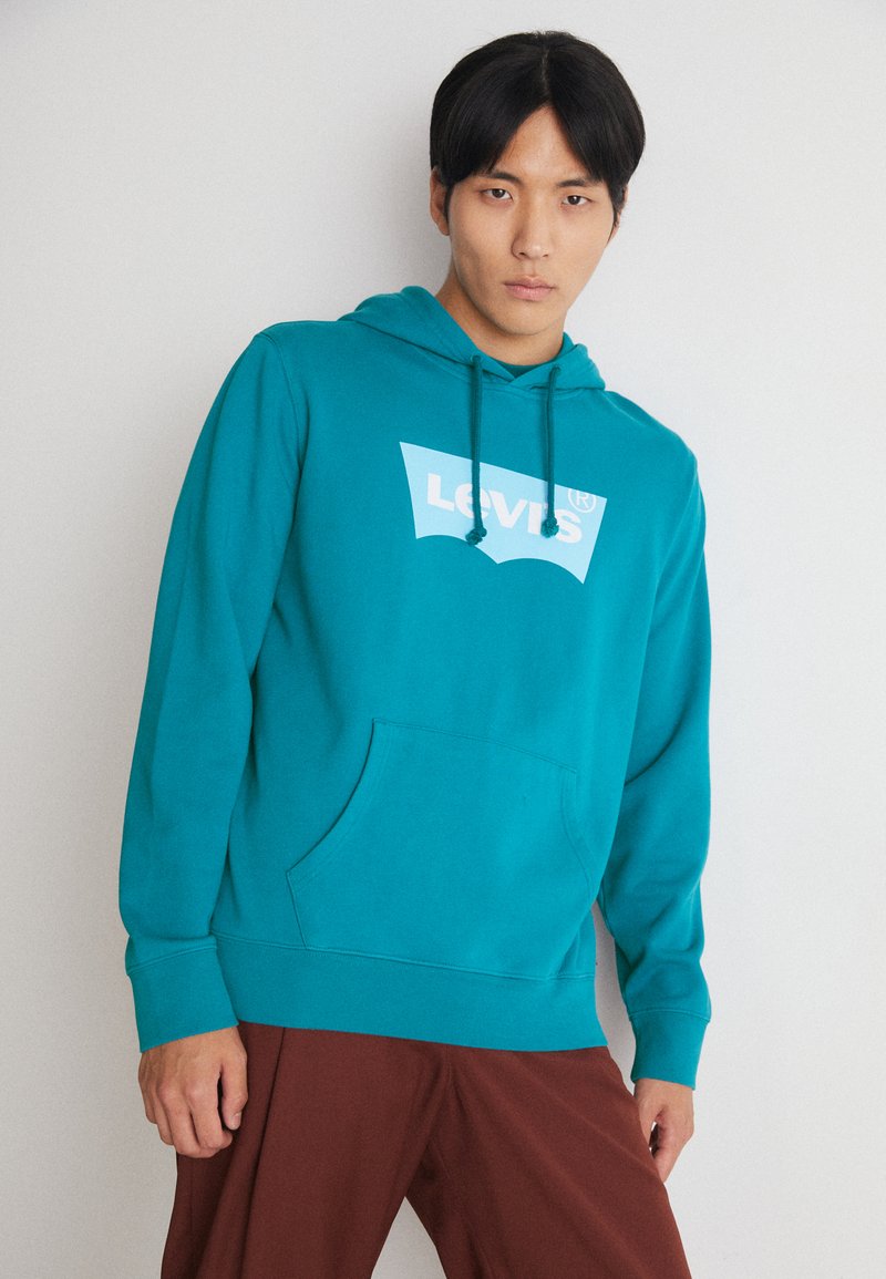 Turquoise hoodie met een witte Levi's logo, trekkoord en een voorkantzak. Model draagt bruine broek met een gladde stofstructuur.