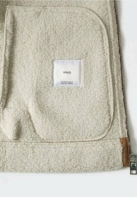 Lys beige fleecejakke med en struktureret overflade, udstyret med lynlås og et firkantet mærke med brandoplysninger øverst til venstre.