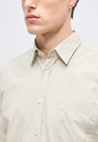 Άνδρας με ανοιχτόχρωμο δέρμα που φοράει μπεζ πουκάμισο με κουμπιά, γιακά και τσέπη στο στήθος, κοιτάζει ελαφρώς προς τα αριστερά σε απλό λευκό φόντο.