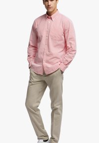 Camicia a righe rosse e bianche con colletto a punta, maniche lunghe e taschino sul petto, abbinata a pantaloni beige e sneaker bianche.