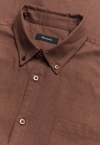 Camicia marrone con bottoni e colletto, realizzata in un tessuto testurizzato, con taschino sul petto e bottoni in tonalità dorata con accenti neri. Etichetta del marchio visibile.