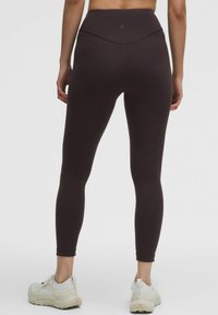 Woman standing facing away wearing dark brown high-waisted leggings and white athletic shoes against a plain gray background.