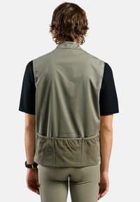 Olivgrüner Weste aus leichtem, glattem Stoff, mit einer Rückentasche und Perforierungen, kombiniert mit einem schwarzen Kurzarmhemd.