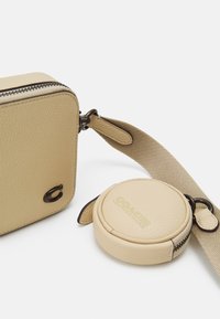 Beige strukturerad lädercrossbodyväska med ett logotyp i guldfärgad hårdvara. Inkluderar en matchande rund myntficka, båda med dragkedjestängning.