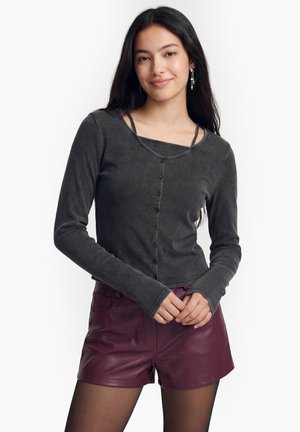 Ung kvinde med langt mørkt hår iført en mørkegrå langærmet top med knapper og bordeauxfarvede lædershorts, smilende med hænderne foldet.