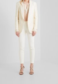 Jasnobeżowy, teksturowany blezer z satynowym wykończeniem, zestawiony z przejrzystą, wzorzystą bluzką oraz dopasowanymi białymi spodniami; minimalistyczne, brązowe sandały na obcasie z cienkimi paskami.