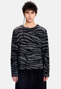 Pull noir et gris flou avec un motif à rayures zèbre, col rond et manches longues, associé à un pantalon foncé.