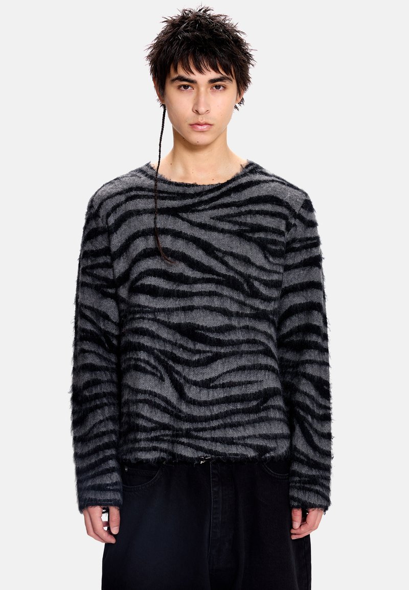 Pull noir et gris flou avec un motif à rayures zèbre, col rond et manches longues, associé à un pantalon foncé.