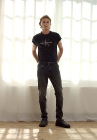 Camiseta de algodón negra con el logo "Calvin Klein Jeans", jeans oscuros y zapatillas negras. El modelo se encuentra de pie frente a un fondo de cortinas transparencias.