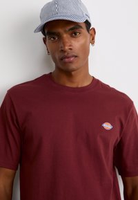 T-shirt in cotone color borgogna con scollatura rotonda e maniche corte. Presenta un piccolo logo blu e arancione sul petto. Cappellino a strisce con visiera curvata.