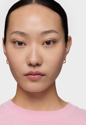 Giovane donna con pelle liscia, che indossa piccoli orecchini a cerchio in oro e un maglione rosa chiaro lavorato a maglia, rivolta verso l'obiettivo con uno sfondo semplice.