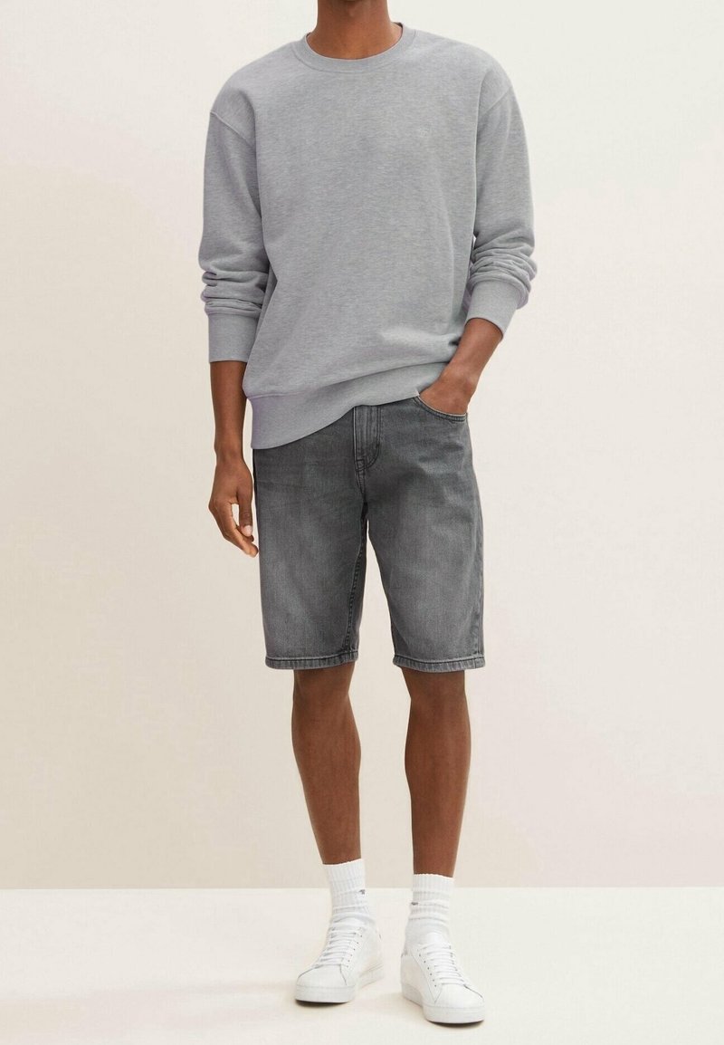 Sudadera gris con puños acanalados, combinada con pantalones cortos de mezclilla gris oscuro. Modelo llevando zapatillas blancas y calcetines tobilleros. Atuendo simple y casual.