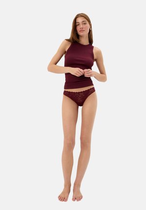 Jeune femme debout pieds nus portant un haut sans manches bordeaux et une lingerie en dentelle assortie, devant un fond clair uni.