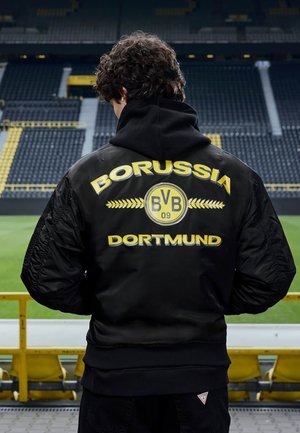 Persona con pelo rizado, con chaqueta negra del Borussia Dortmund, de espaldas a las gradas vacías del estadio y al campo de fútbol verde.