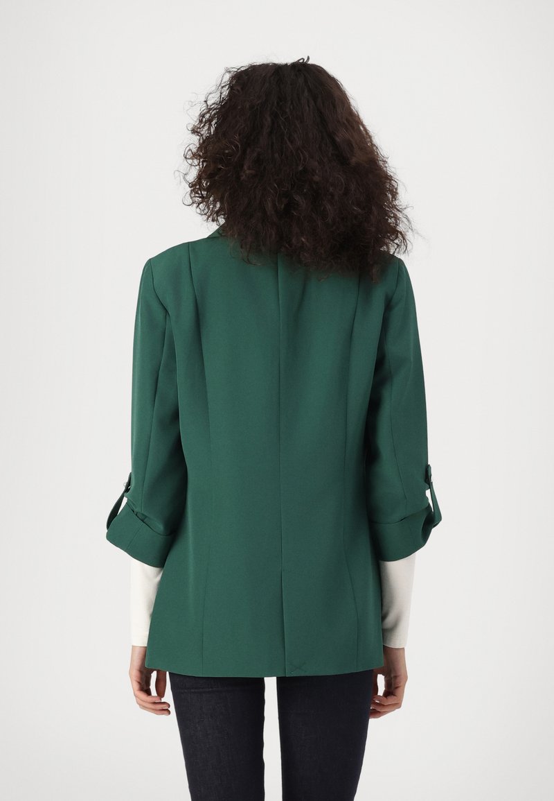 Žena s kovrčavom kosom u tamnozelenom sakou s podvrnutim rukavima stoji okrenuta leđima na jednobojnoj pozadini.